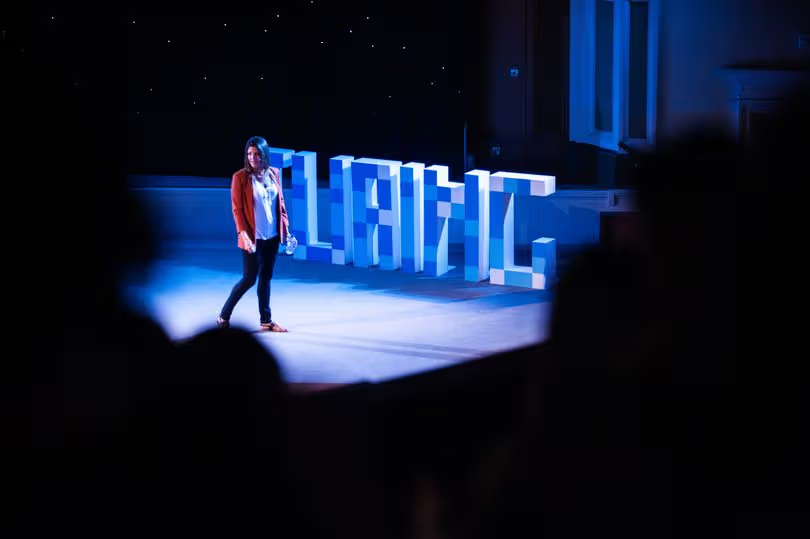 A woman walking on a stage in front of large blue letters spelling TURING