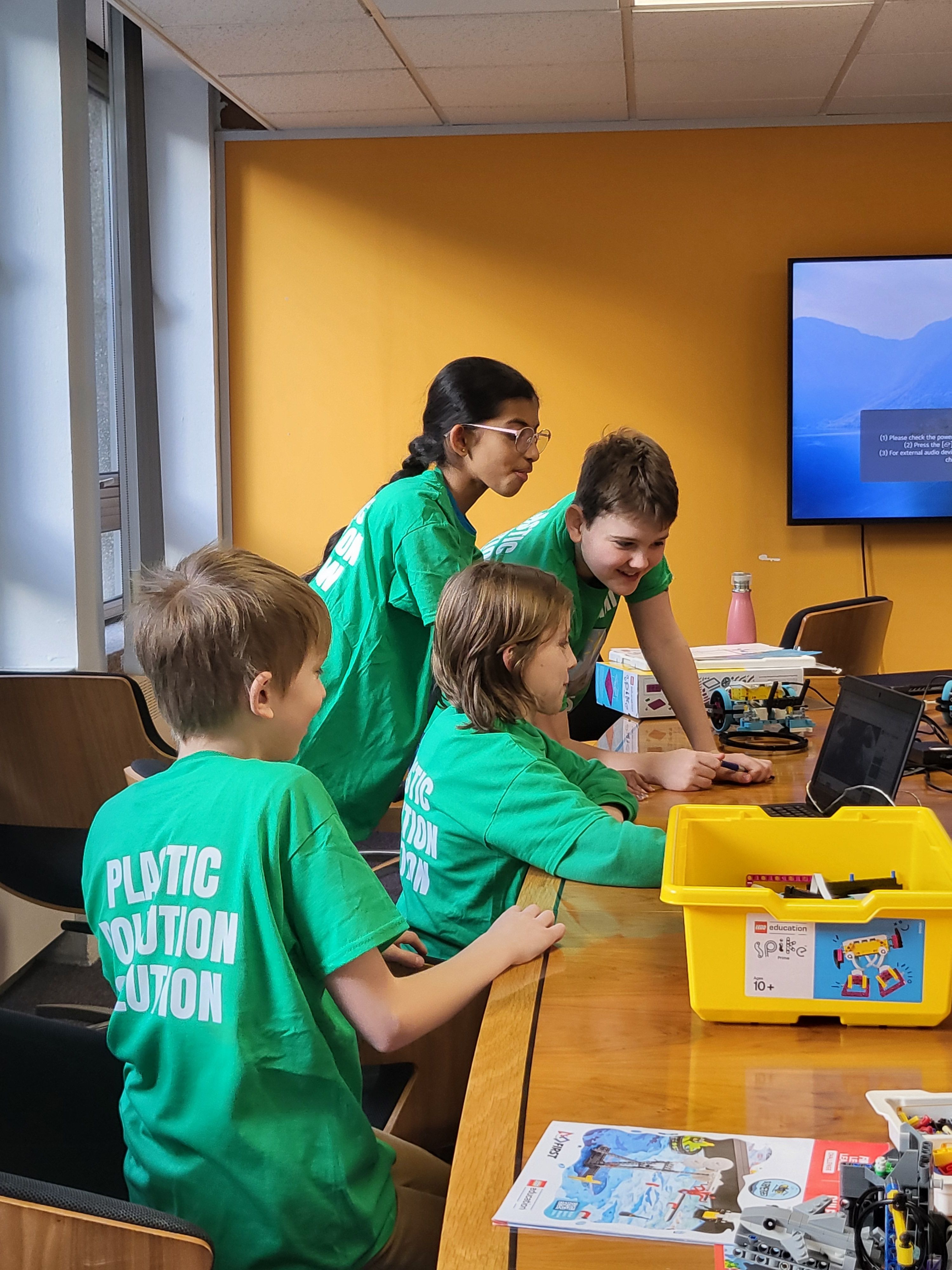 Four children wearing t-shirts that read 'Plastic Pollution Solution' looking at a laptop surrounded by LEGO