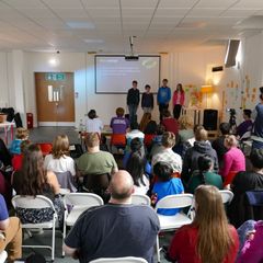 Four young people presenting a slide in front of an audience of about 30 listening adults