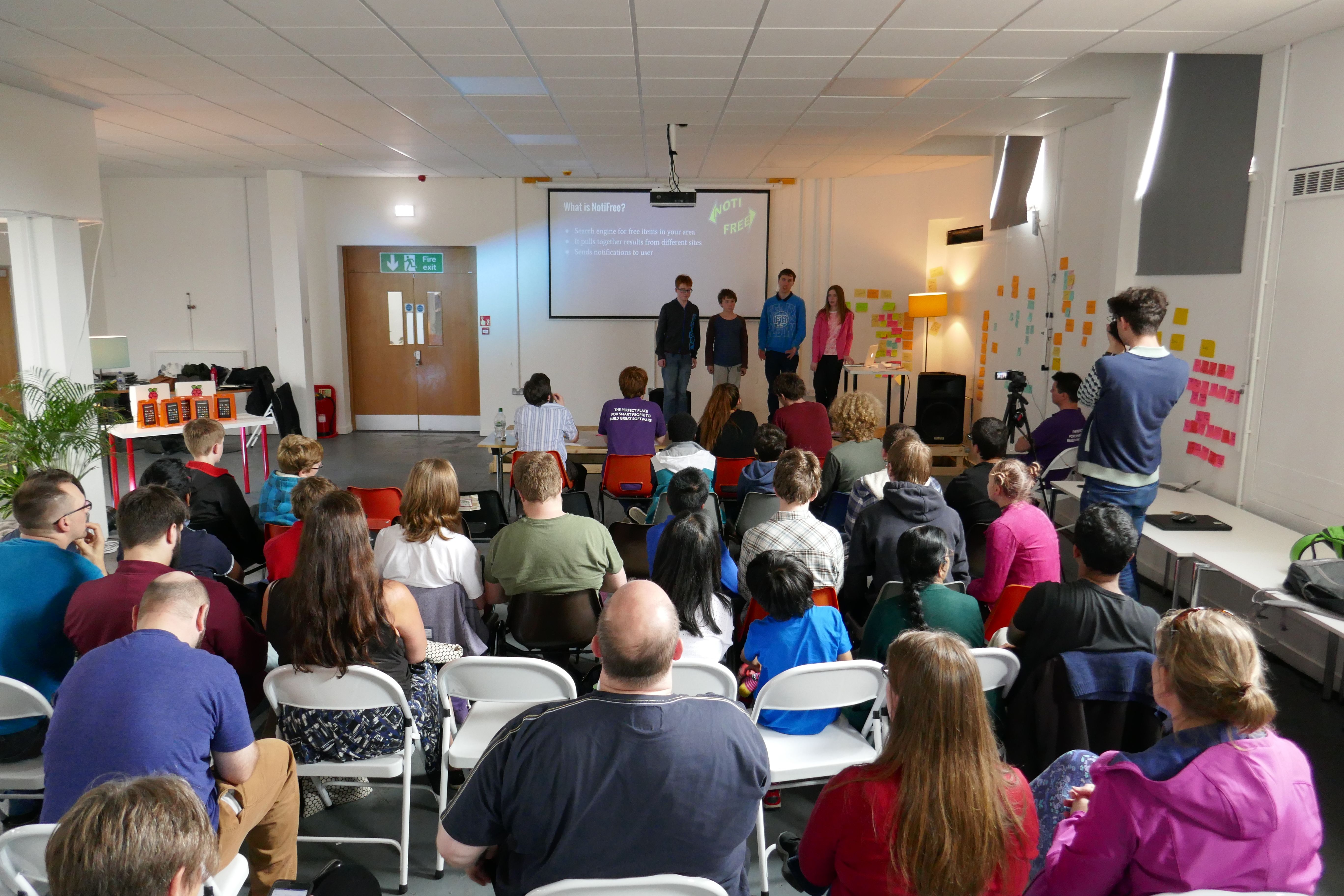 Four young people presenting a slide in front of an audience of about 30 listening adults