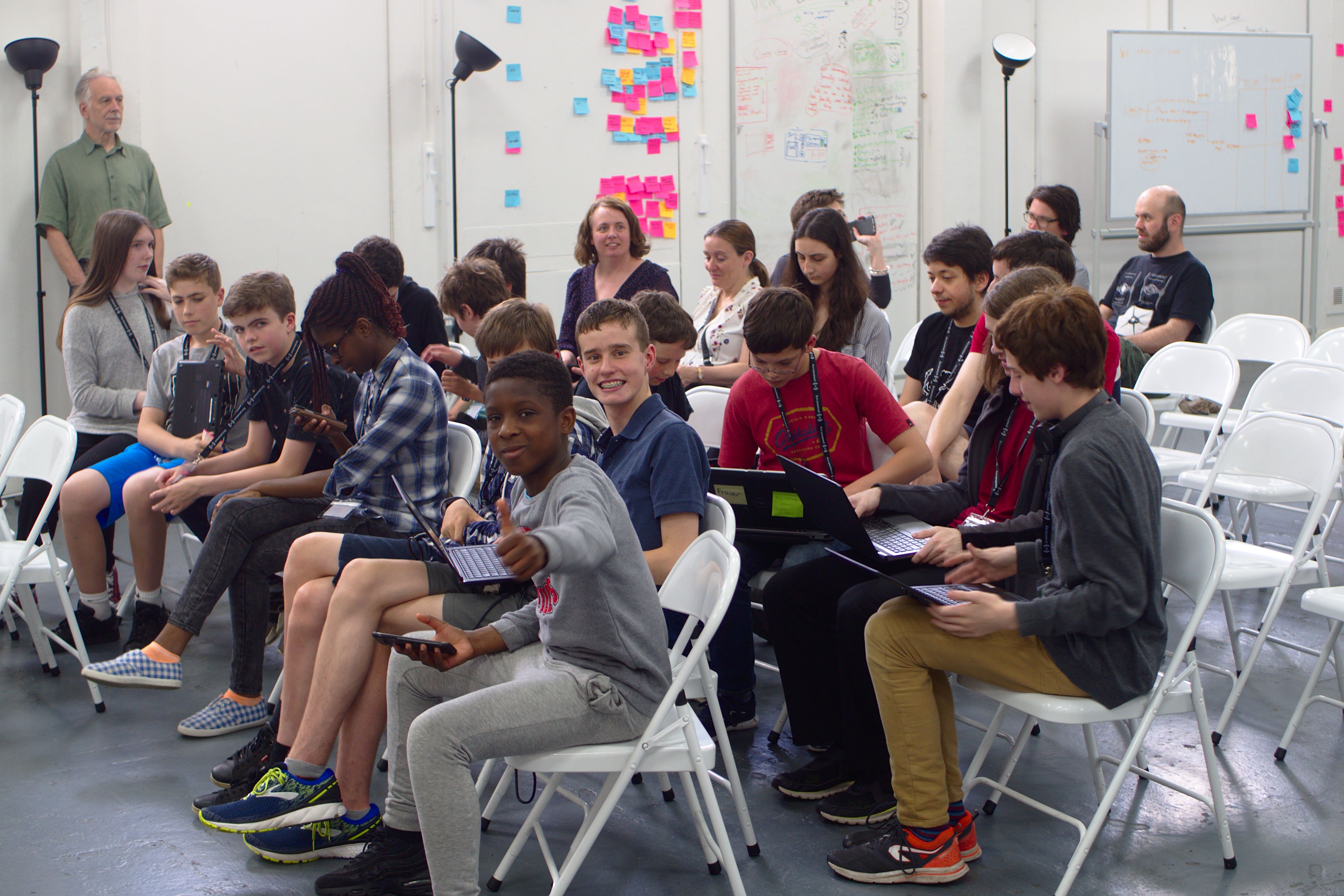About 20 young people sitting on several rows of chairs, with some looking at a laptop together and one giving a thumbs up to the camera
