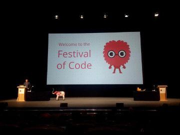 A presenter on a stage with a slide that says "Welcome to the Festival of Code" behind them